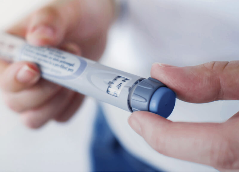 A close-up of hands holding a medical injection pen, with fingers turning the dose dial to set the correct amount.