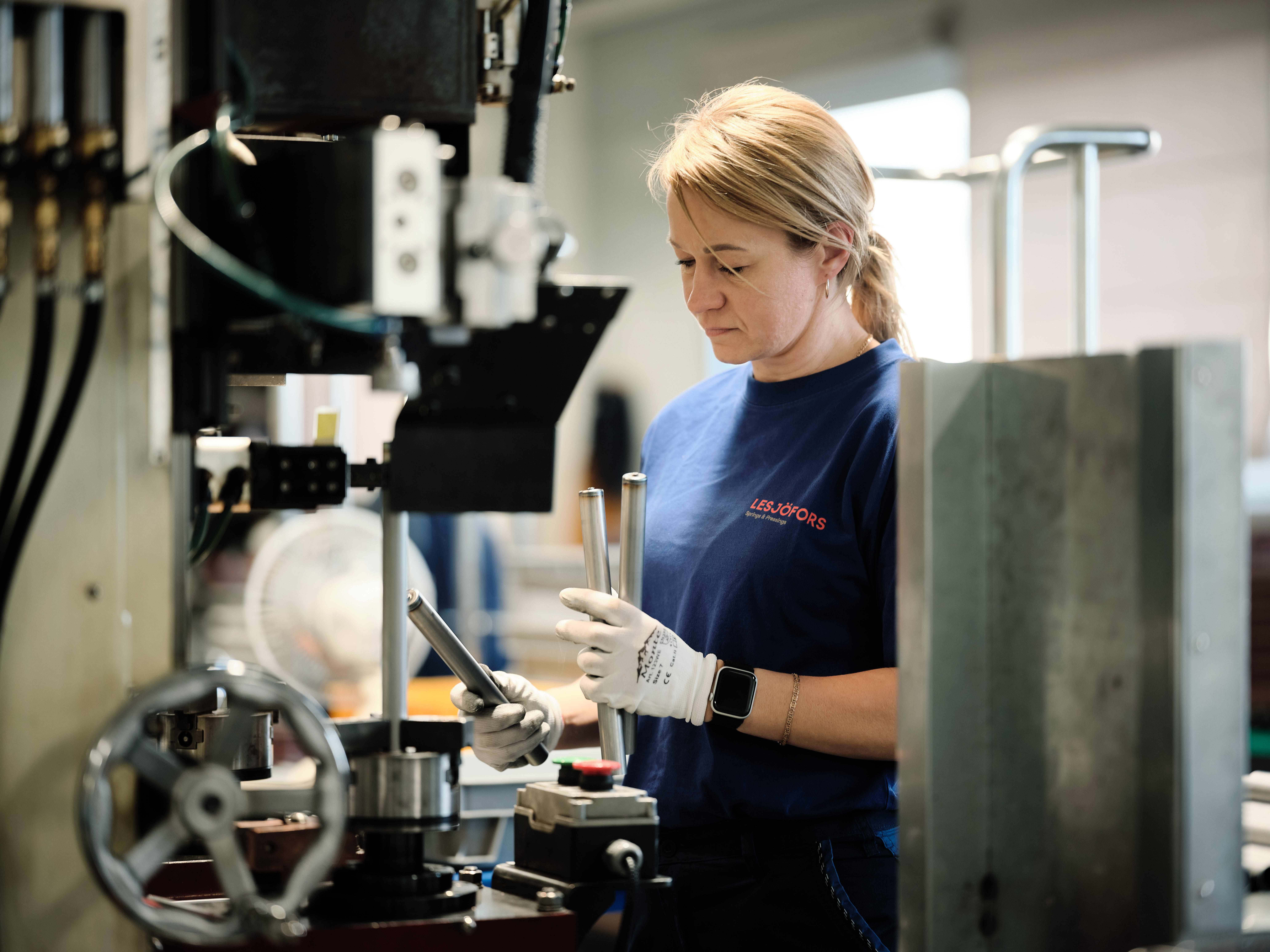 A woman in a blue shirt and gloves works intently with springmachinery in a factory.