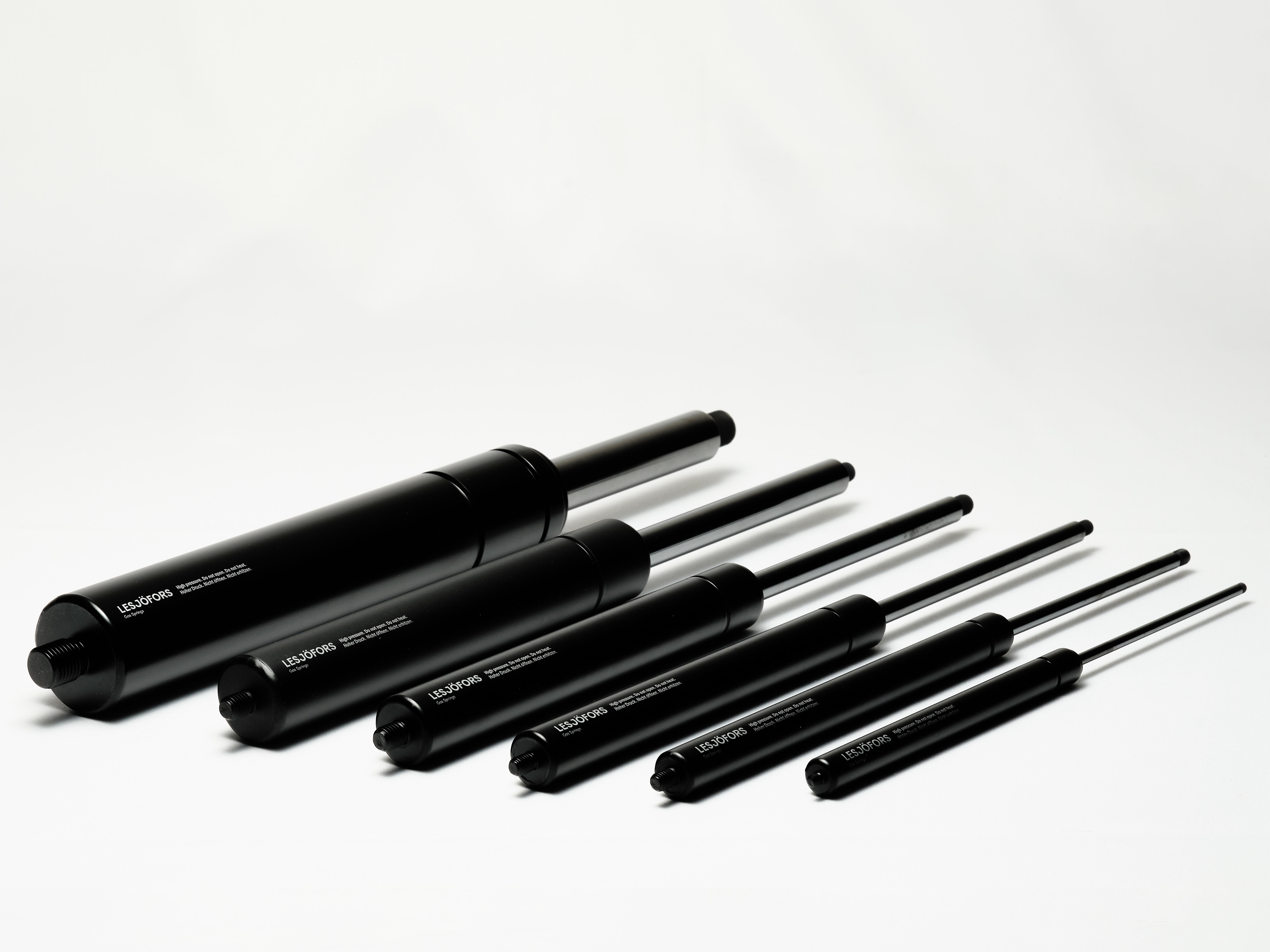 A row of six black and silver dynamic gas springs of varying sizes is arranged on a white surface. The dynamic gas springs are angled to the left, creating a sleek and organized look.