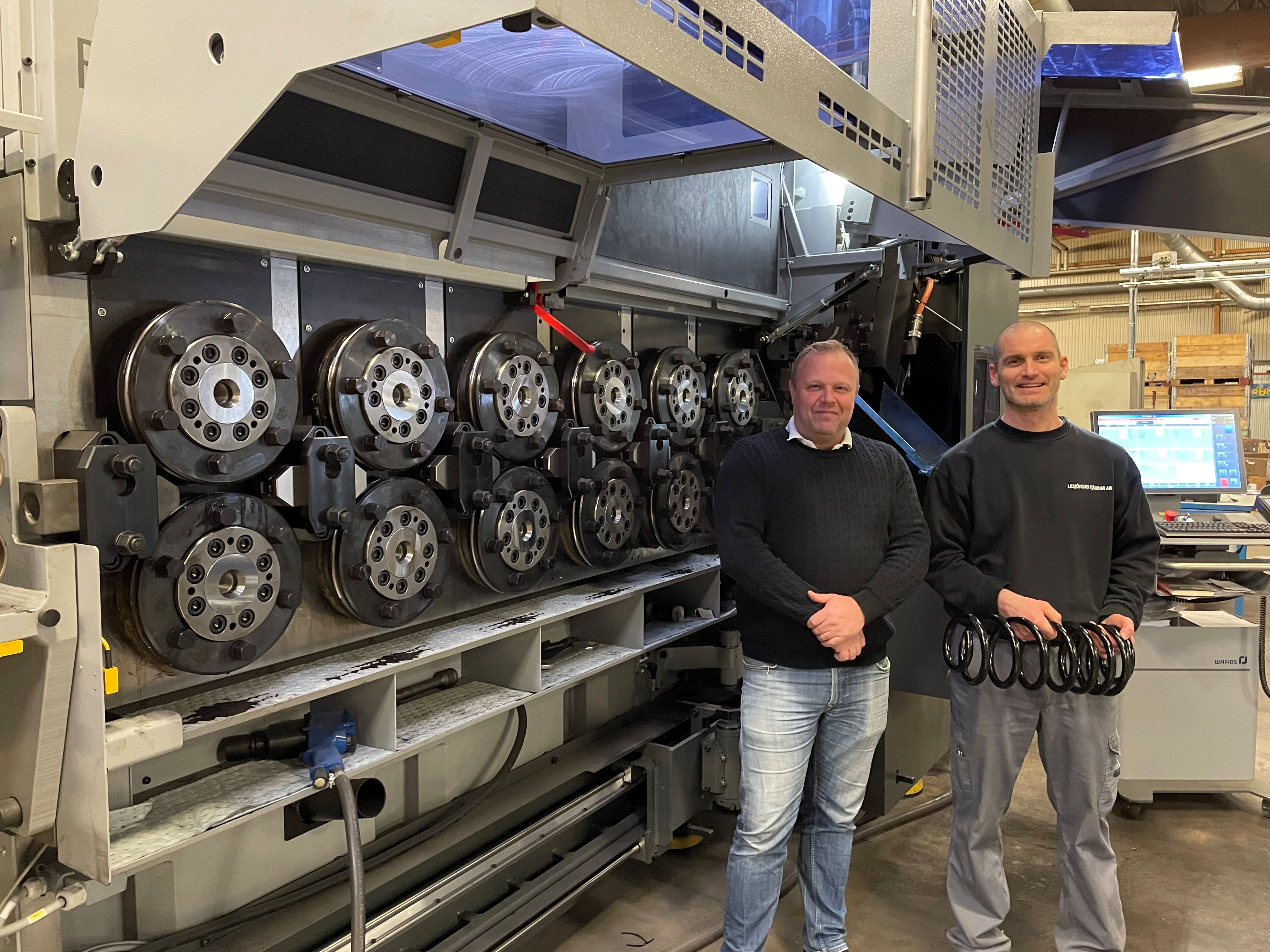 Two people stand smiling in front of a large industrial machine with multiple wheels. The setting appears to be a workspace with a relaxed, casual tone.