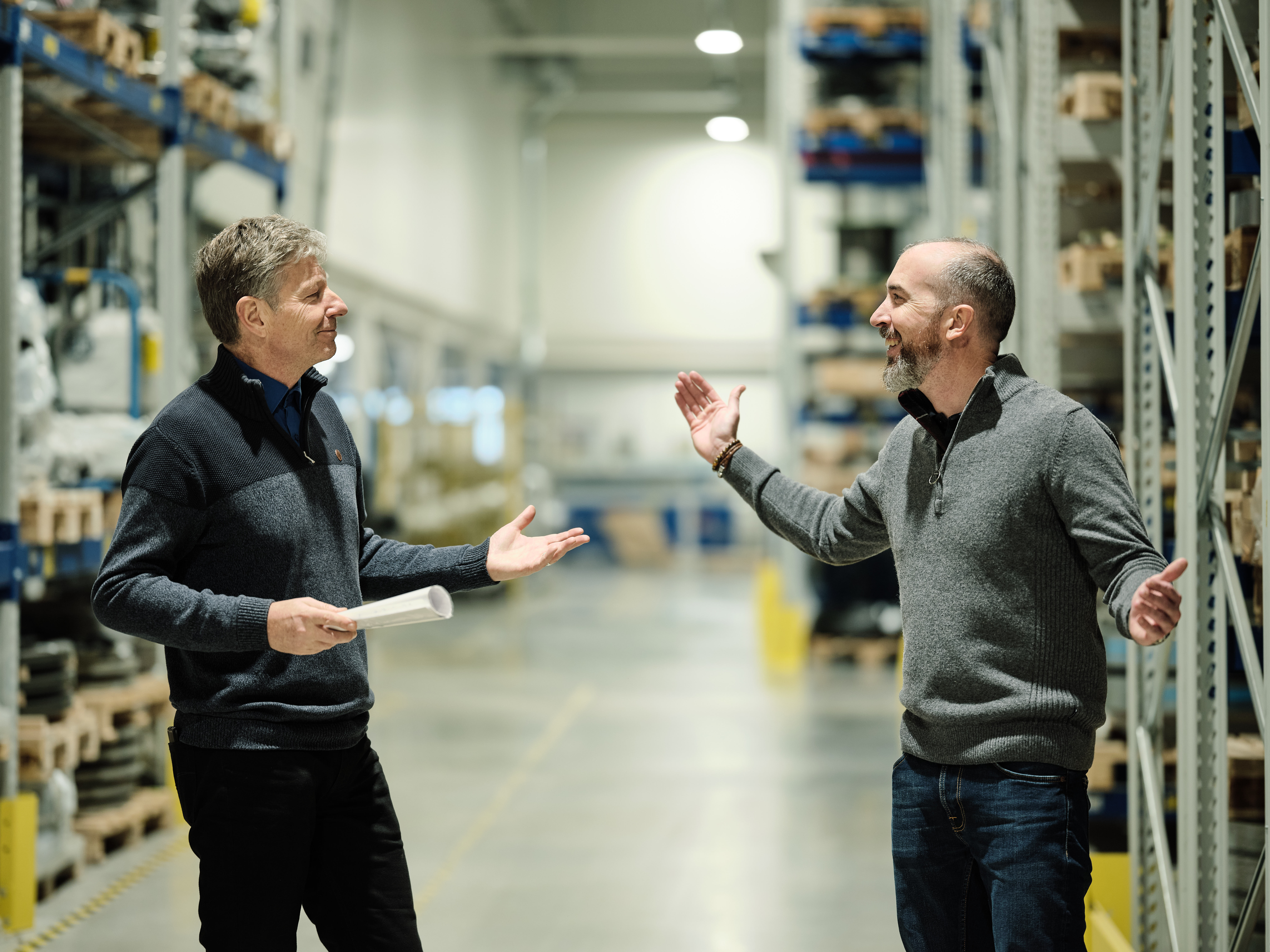 Two men in a warehouse, smiling and gesturing as they talk. Shelves filled with boxes line the walls, under bright industrial lighting.