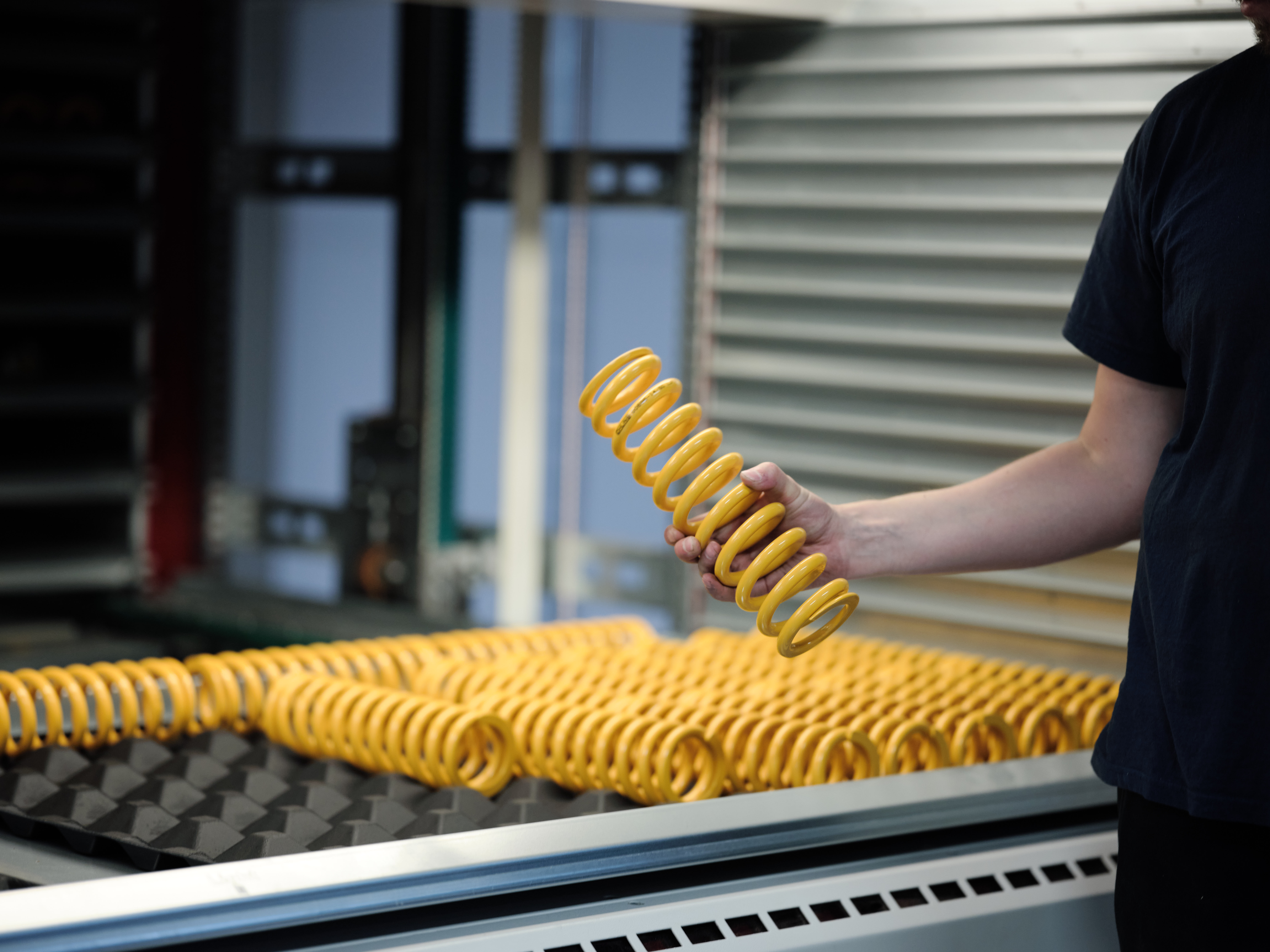 Worker holding a yellow-coated coil spring above a conveyor with multiple identical springs.