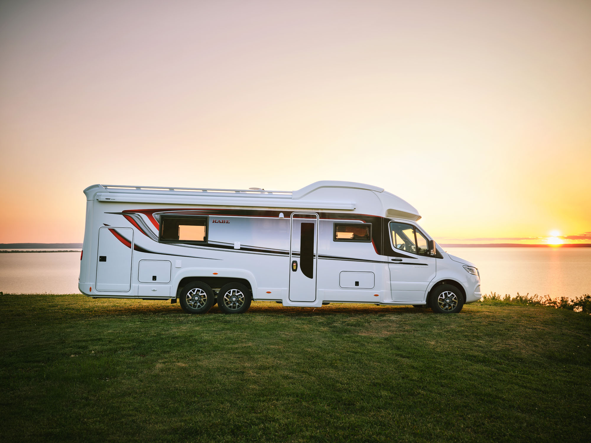 Camper van parked by a serene lakeside at sunset, with a soft orange and pink sky.