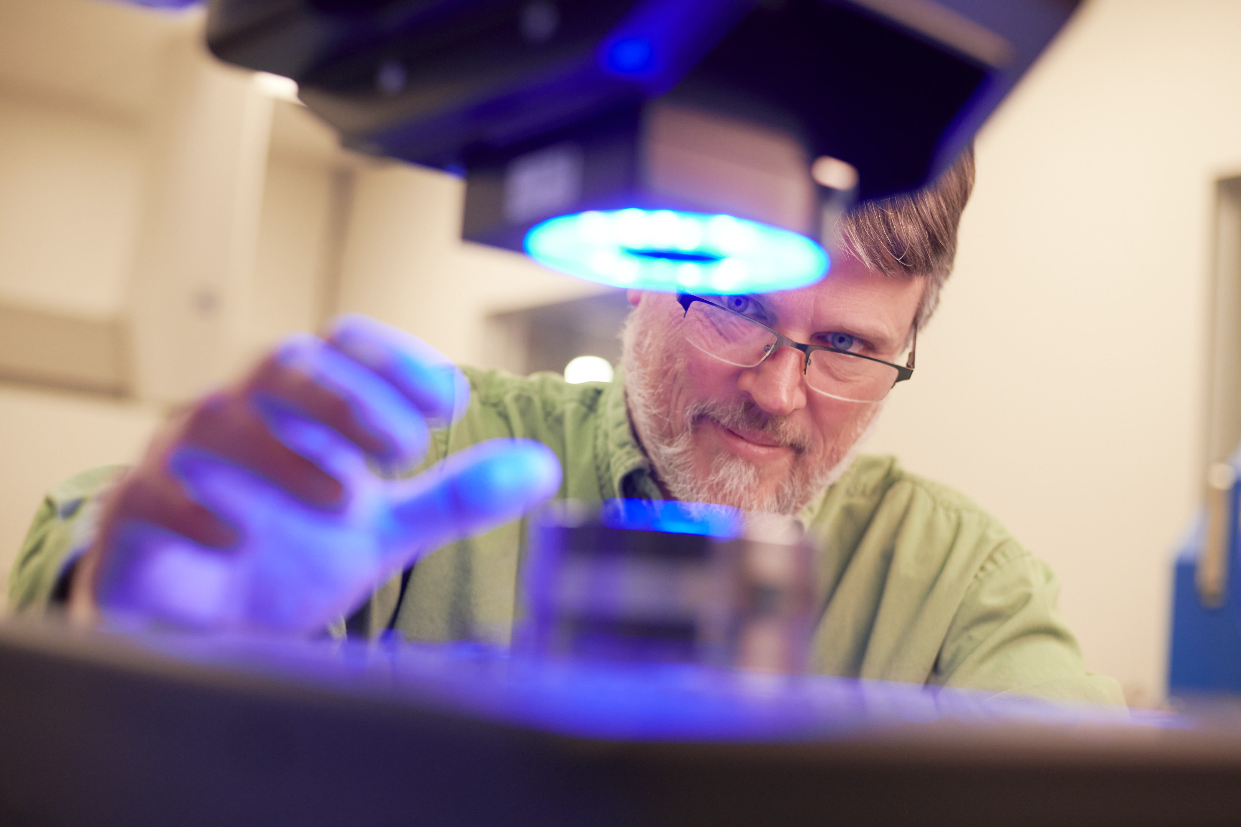 A person in a laboratory is focusing on a microscope with blue light, suggesting some type of production.