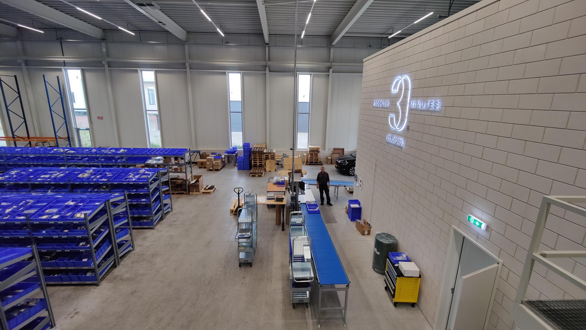 A spacious industrial warehouse with rows of blue crates on the left, workers handling goods on conveyor belts in the center, and a large logo on the right wall.