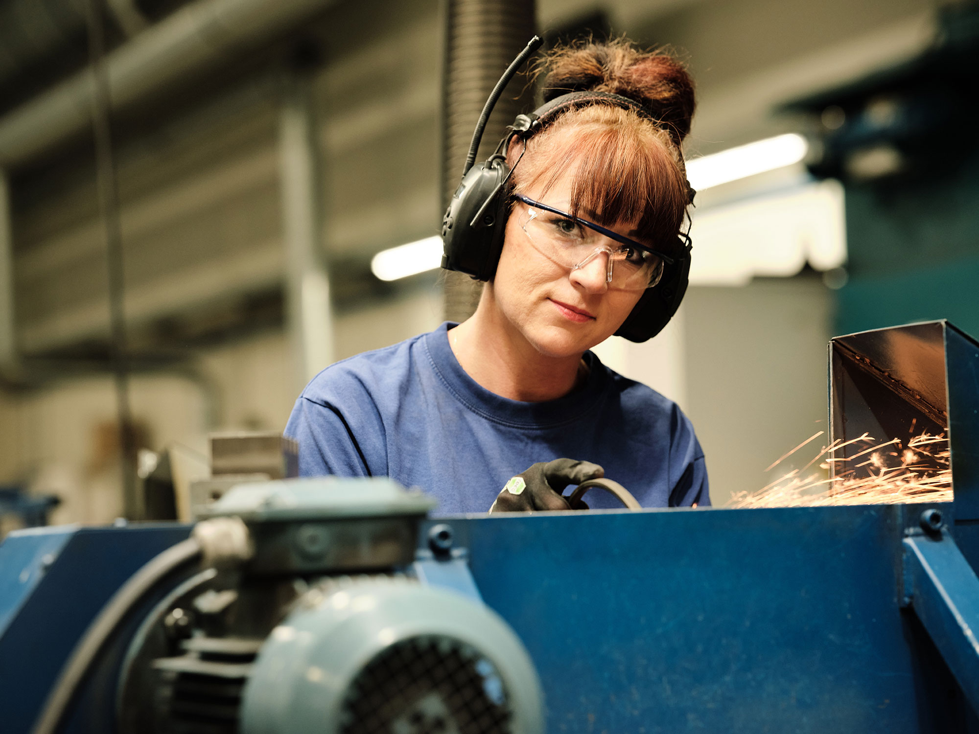 Factory worker operating a machine as sparks fly, wearing safety glasses and ear protection.