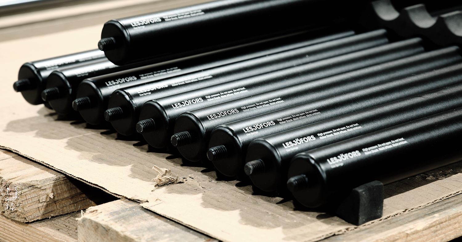 Stacked black gas springs on a wooden pallet, aligned neatly in rows. The shiny black surfaces suggest newness.
