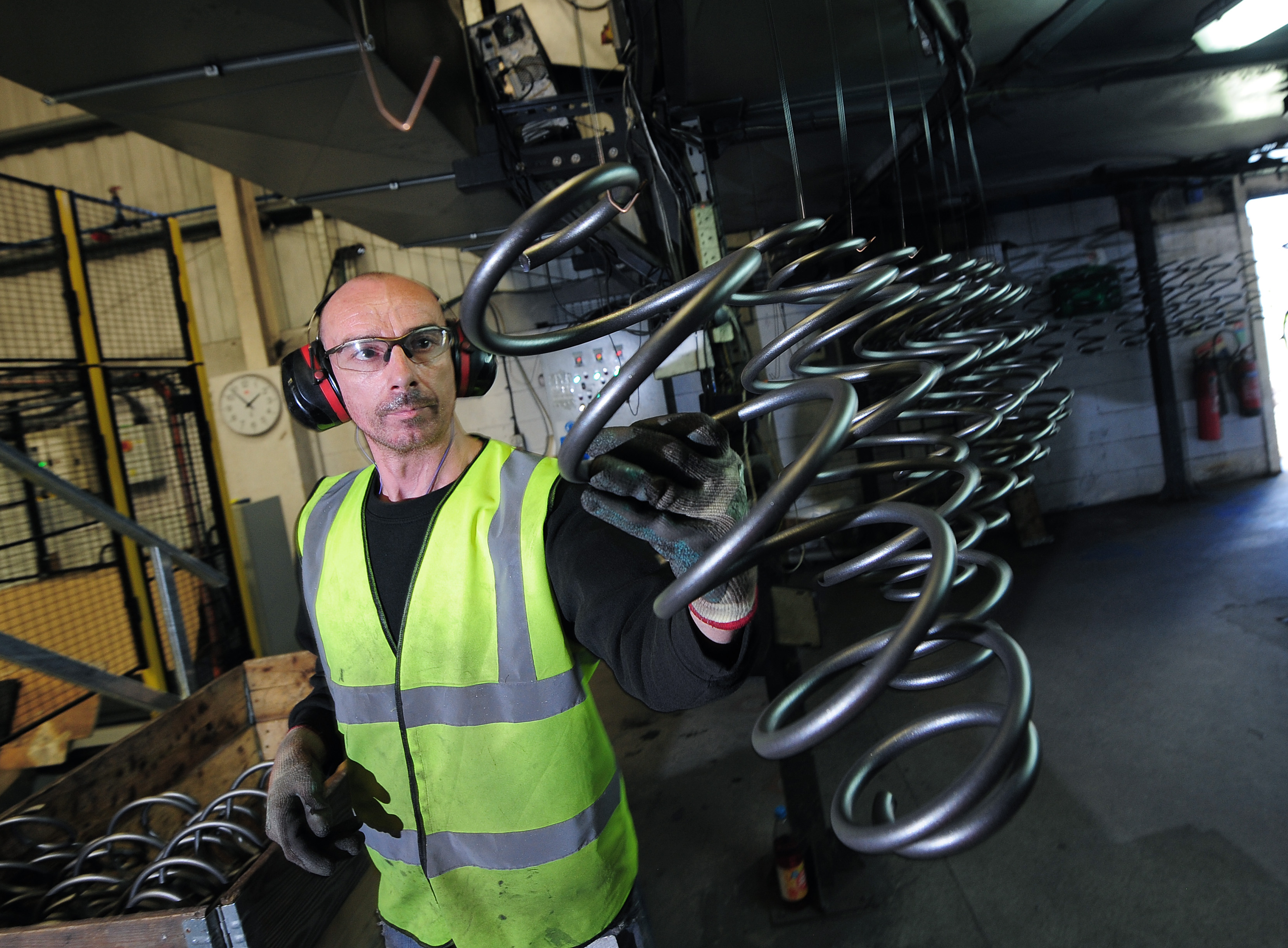 A factory worker wearing safety gear inspects large metal coils with a focused expression.