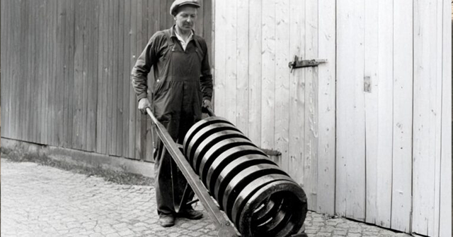 Black-and-white photo of a worker moving a large coil spring on a hand cart.