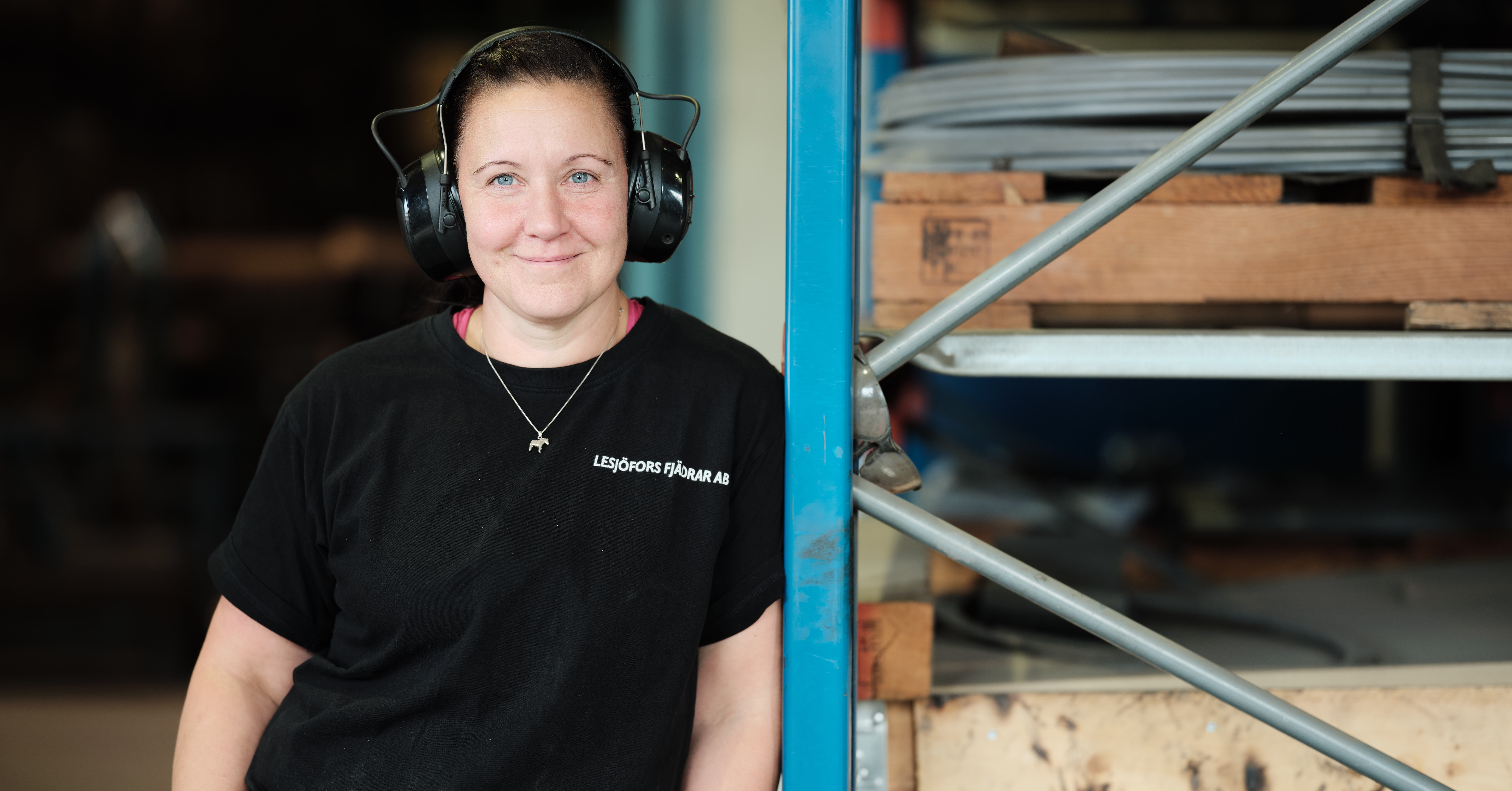 A person with short hair, wearing headphones and a black t-shirt, stands smiling beside industrial shelves in a workshop setting.