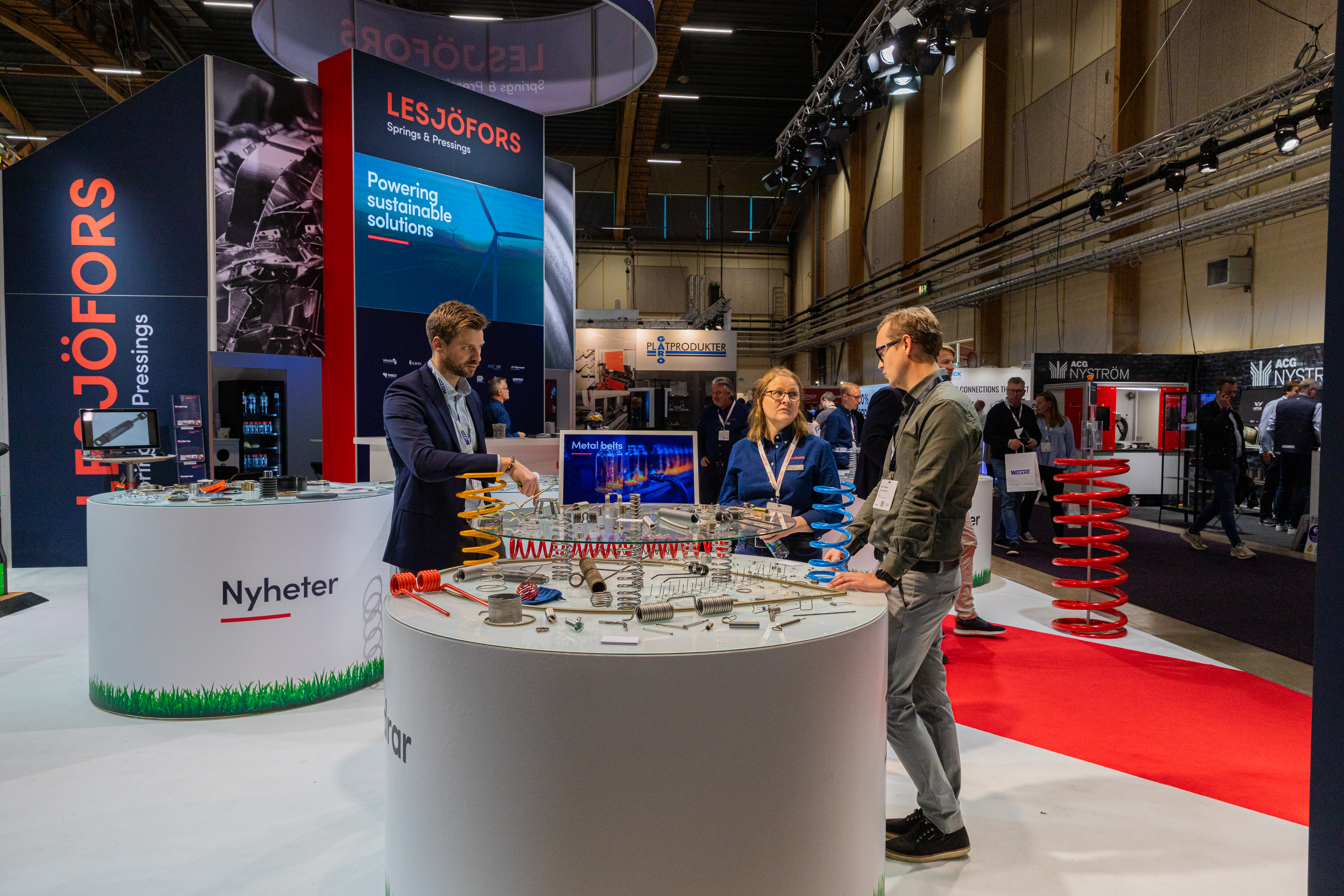 Three people discussing products at a Lesjöfors exhibition stand, with various industrial springs and metal components displayed on a round table.