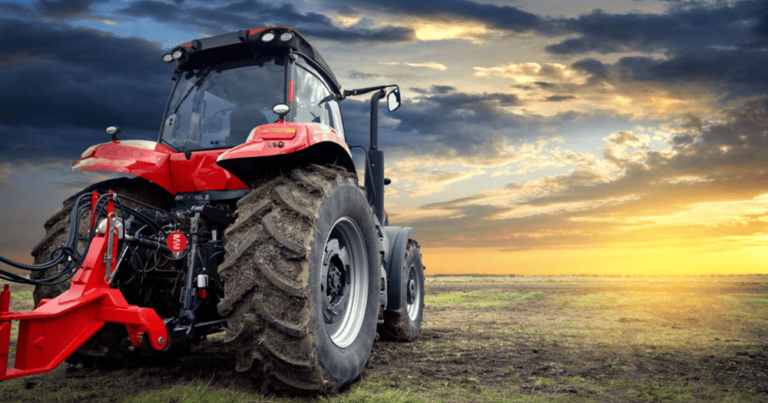 A red tractor with large tires is parked on a grassy field under a dramatic sunset.