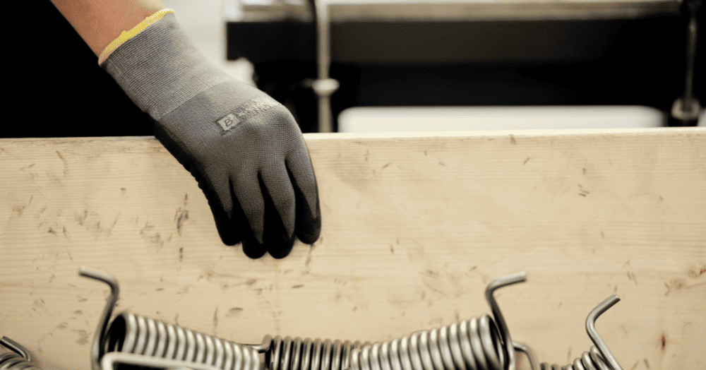 A gloved hand rests on a wooden plank, with a pile of metal springs in the foreground.