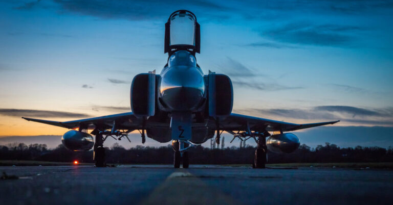 A fighter jet is parked on a runway at dusk, silhouetted against a dramatic blue and orange sky.
