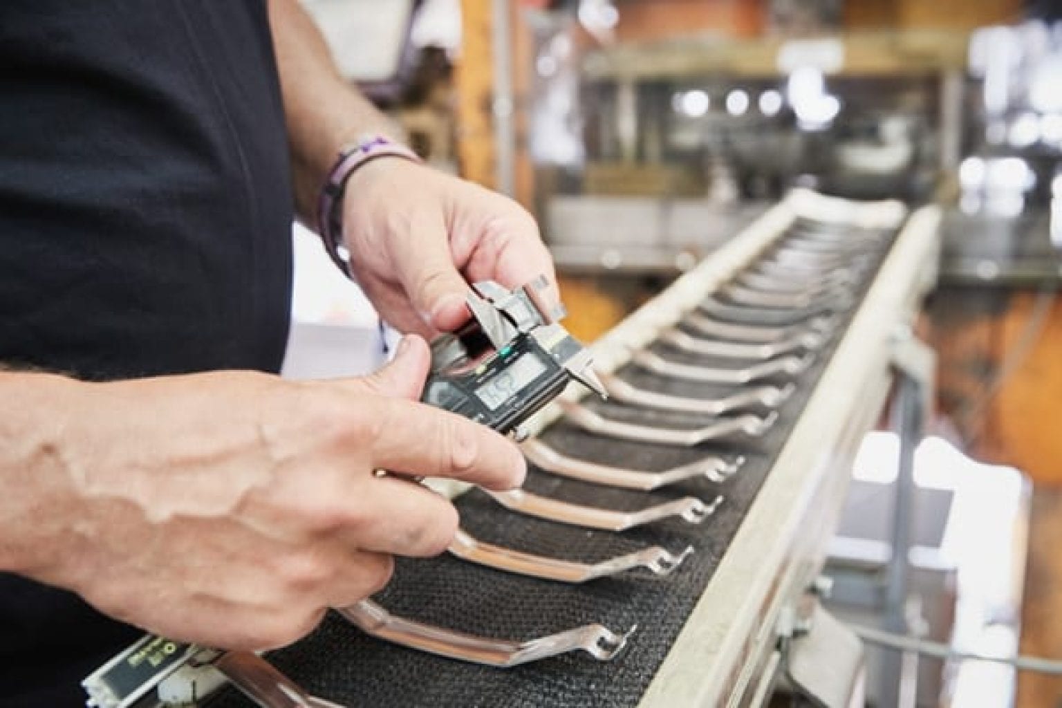 A worker using digital calipers to measure a formed metal part, with multiple identical components lined up on a conveyor belt.