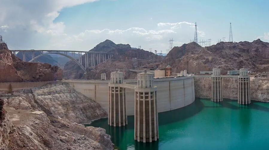 Large concrete dam with water reservoir surrounded by rocky landscape and bridge in background.