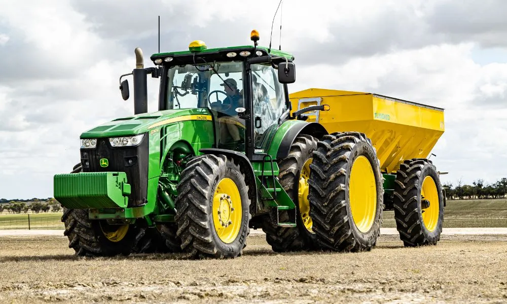 Large green tractor pulling a yellow trailer on farmland under a cloudy sky.