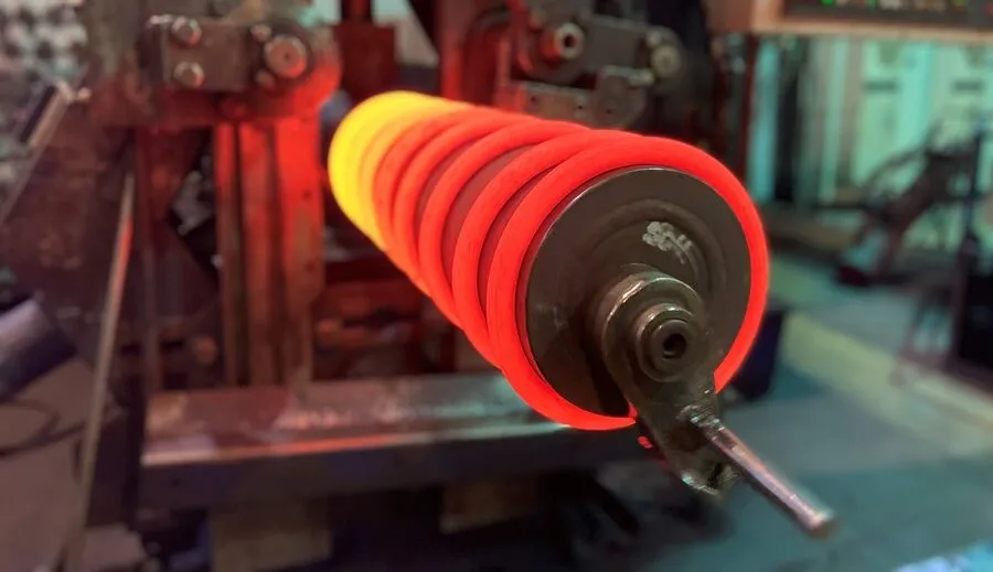 A close-up of a glowing red and orange hot coiled compression spring on a mandrel during the hot coiling manufacturing process.
