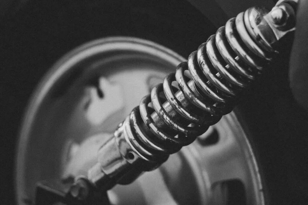 A black and white close-up photograph of a compression spring on a vehicle suspension system, with a wheel rim visible in the background.