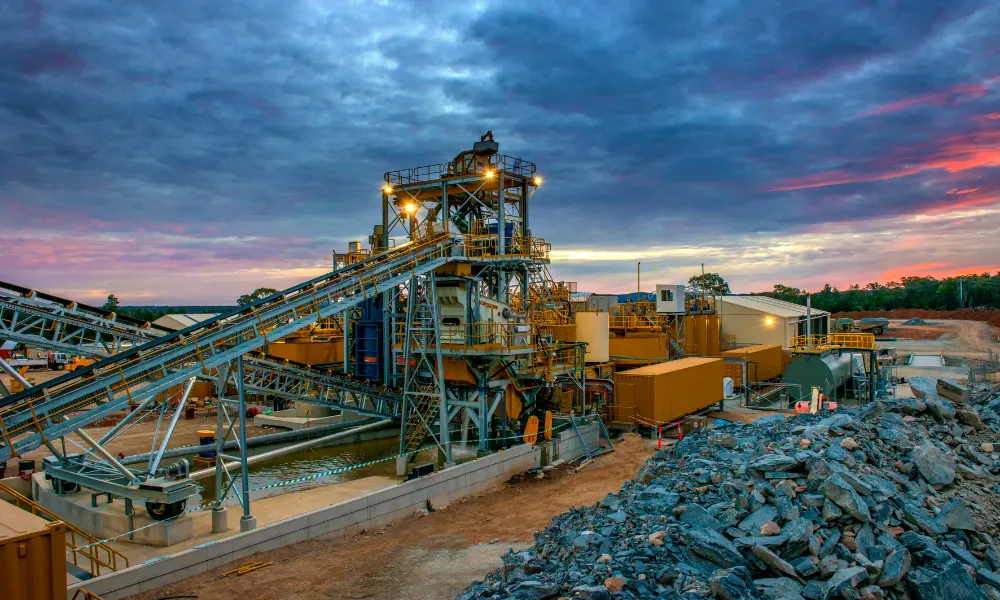 Illuminated mineral processing plant with conveyor systems and stockpiled rock at dusk with a dramatic sunset sky.