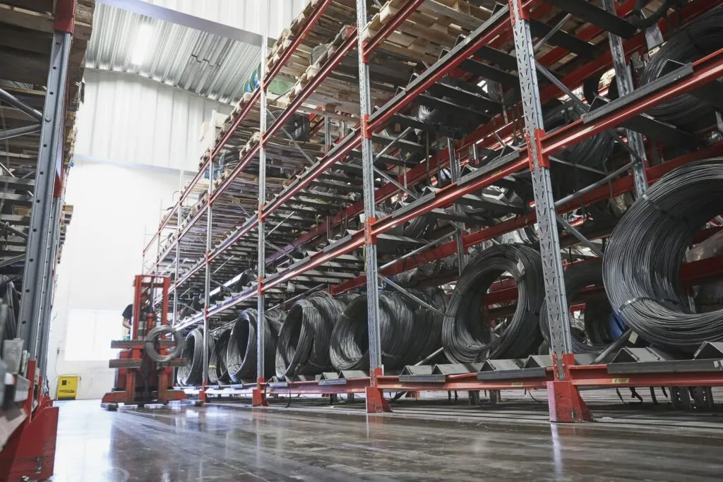 Interior of a large industrial warehouse showing tall red metal racking systems storing multiple coils of steel wire rod stacked across several levels, with a polished concrete floor in the foreground.