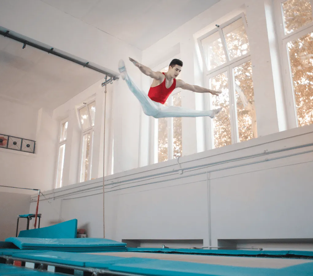 A male trampolinist in a red leotard and white trousers performing a pike jump high above a blue trampoline in an indoor gymnastics gym.