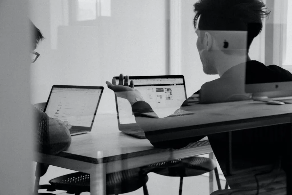 Two people collaborating at a desk with laptops about coil spring design, one gesturing towards the screens during a discussion, photographed in black and white.