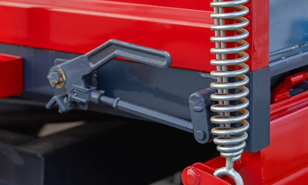 A close-up of a silver tension spring with a hook end mounted vertically on red and grey heavy machinery, alongside a metal latch mechanism.