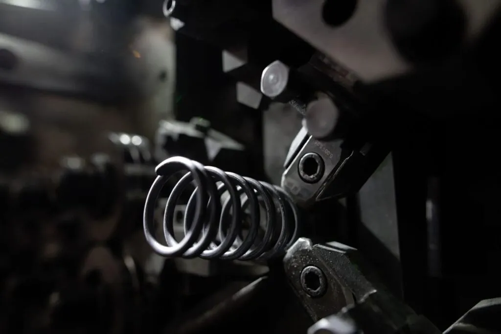 Close-up of a steel coil spring being formed on an industrial coiling machine, with mechanical components and tooling visible in the dark, blurred background.