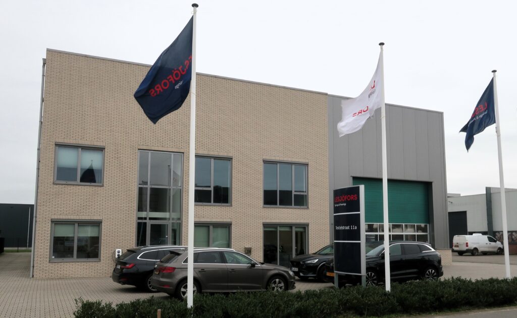 A modern beige office building with large windows, three flags in front—two dark, one white—and several parked cars, set against a gray sky.