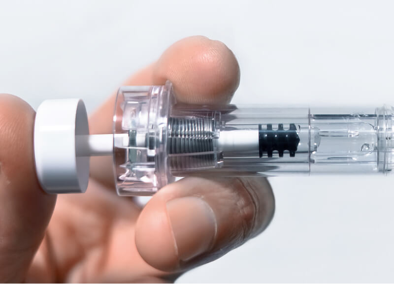 A hand holds a transparent syringe-like device with a visible spring mechanism inside. The image conveys a clinical, precise, and clean appearance.