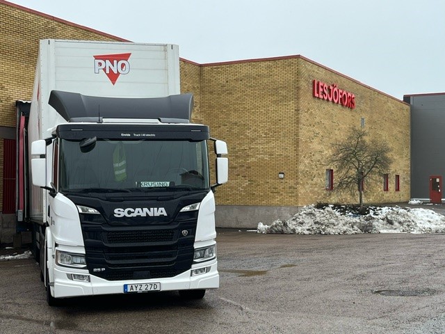 A white and black Scania truck is parked outside a brown brick warehouse on a cloudy day. Snow is piled near the building.