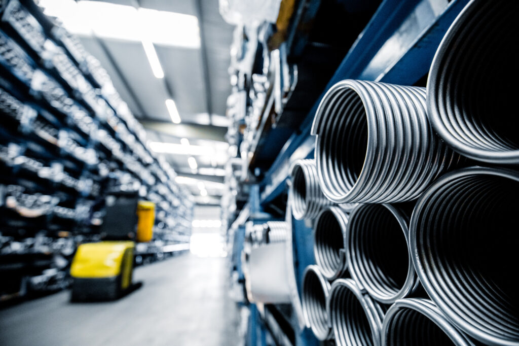 A brightly lit industrial warehouse aisle with stacked metal pipes on the right and a yellow forklift in the distance.