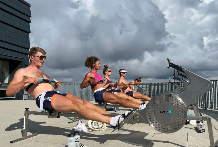 Four people exercise intensely on rowing machines outdoors. They're focused, under a dramatic cloudy sky.