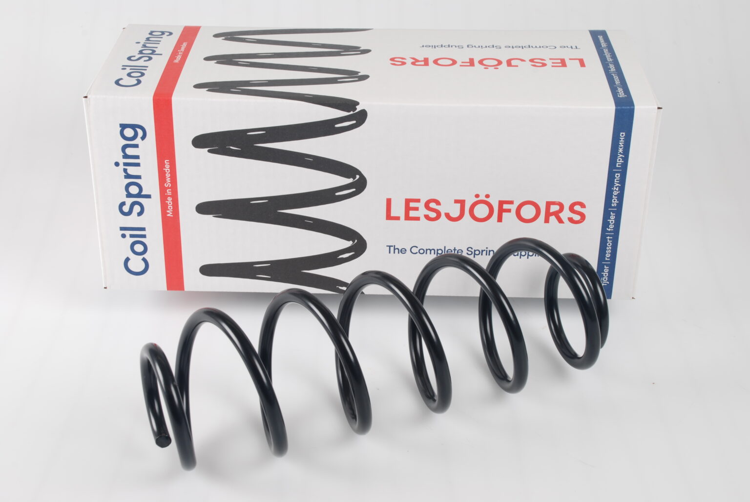 Close-up of a black coil spring in front of a white box labeled "Lesjöfors" with red and blue accents. The spring is tilted and casts a shadow.