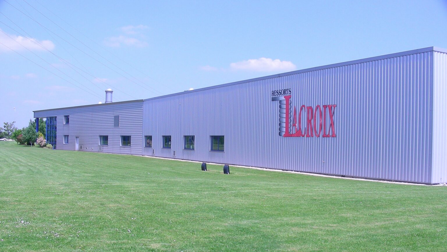 A long, white industrial building with large red letters on the side. It is set against a bright blue sky and surrounded by green grass.