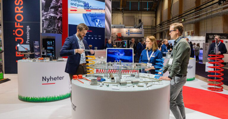 Three people stand around a circular booth display at an industrial trade show. Various metal springs and parts are showcased. The setting is bright and professional.