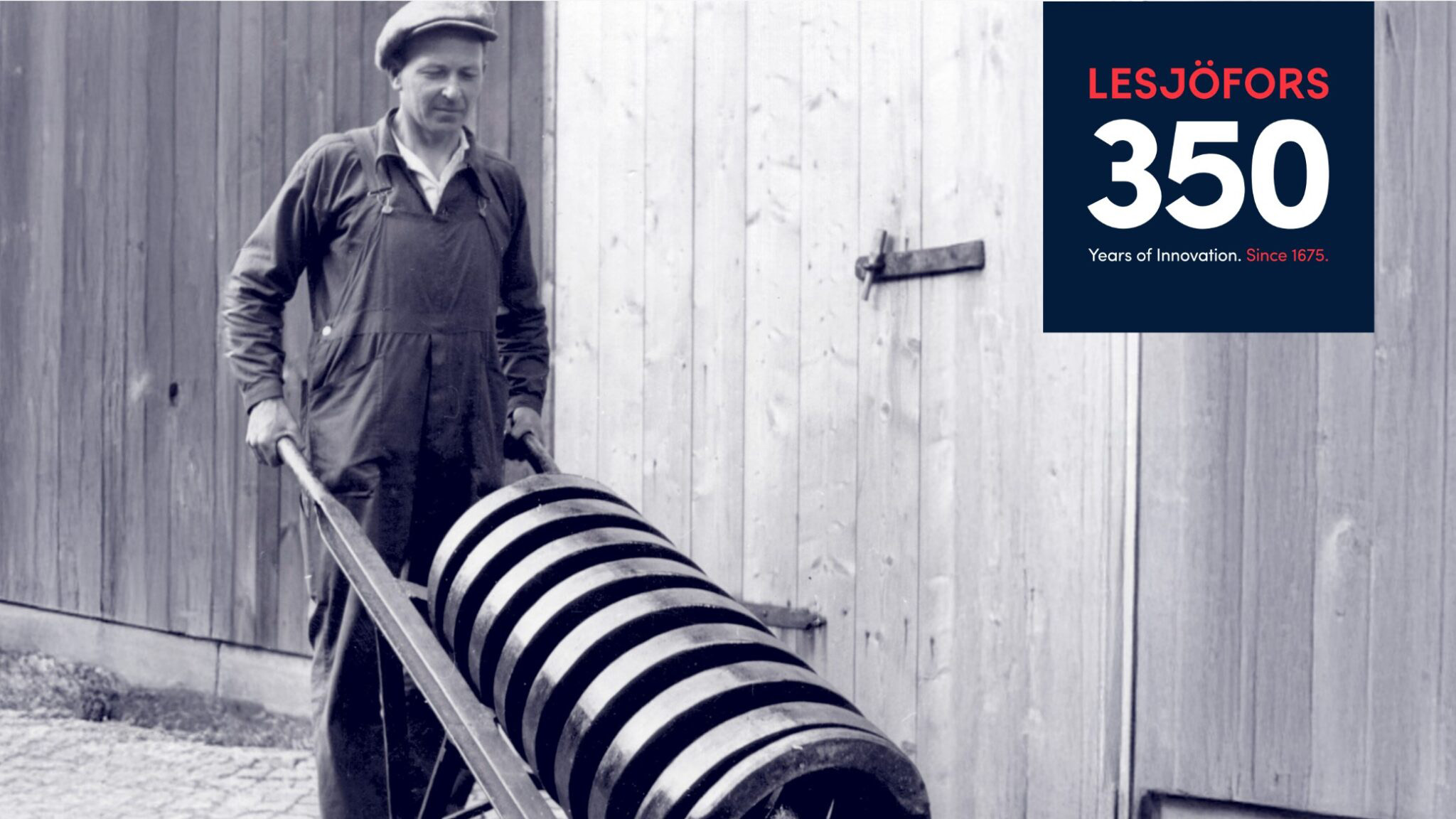 A man in overalls and a cap pushes a cart with large coiled springs on a cobblestone path. A wooden barn door is visible. Text: "Lesjöfors 350 Years of Innovation. Since 1675."