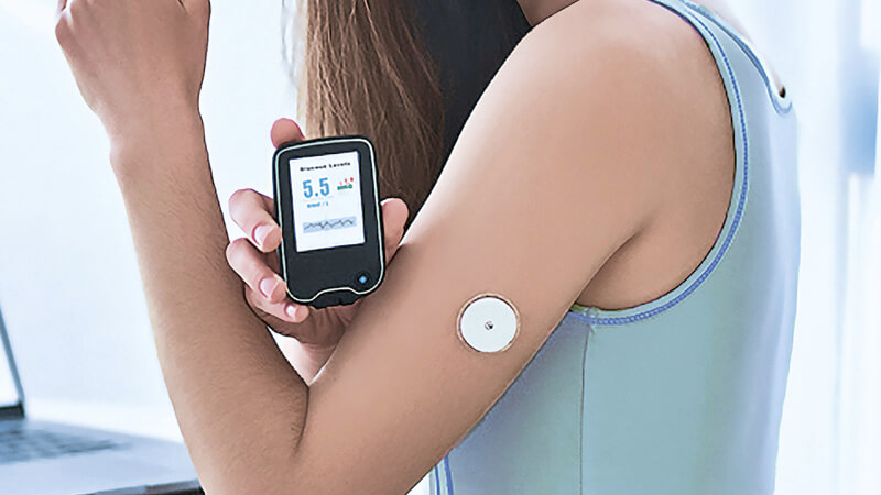 A woman holds a glucose monitor displaying "5.5" next to a sensor on her arm, hinting at diabetes management. She wears a light blue tank top.