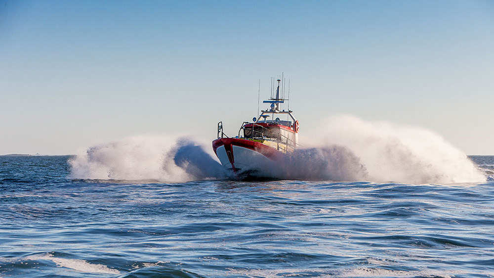 A red and white rescue boat speeds through the ocean, creating large waves and spray against a clear blue sky, conveying urgency and action.