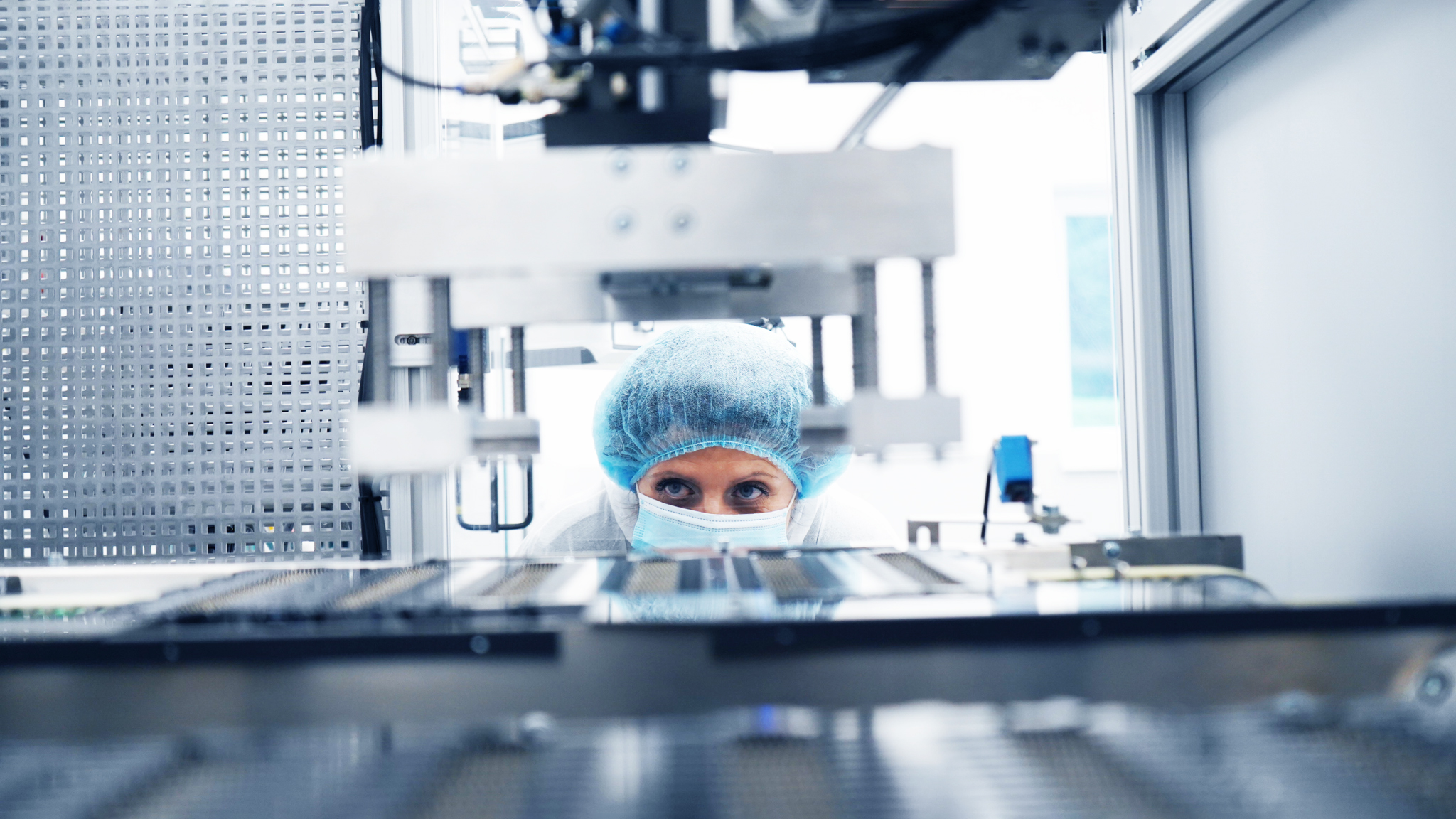 A person in a blue hairnet and face mask focuses intently on machinery in a sterile lab, conveying precision and concentration in a high-tech setting.