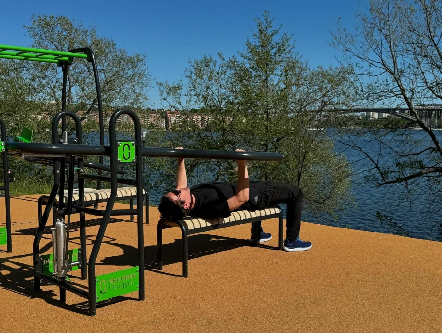 A person exercises on outdoor gym equipment by a river, surrounded by trees under a clear blue sky.