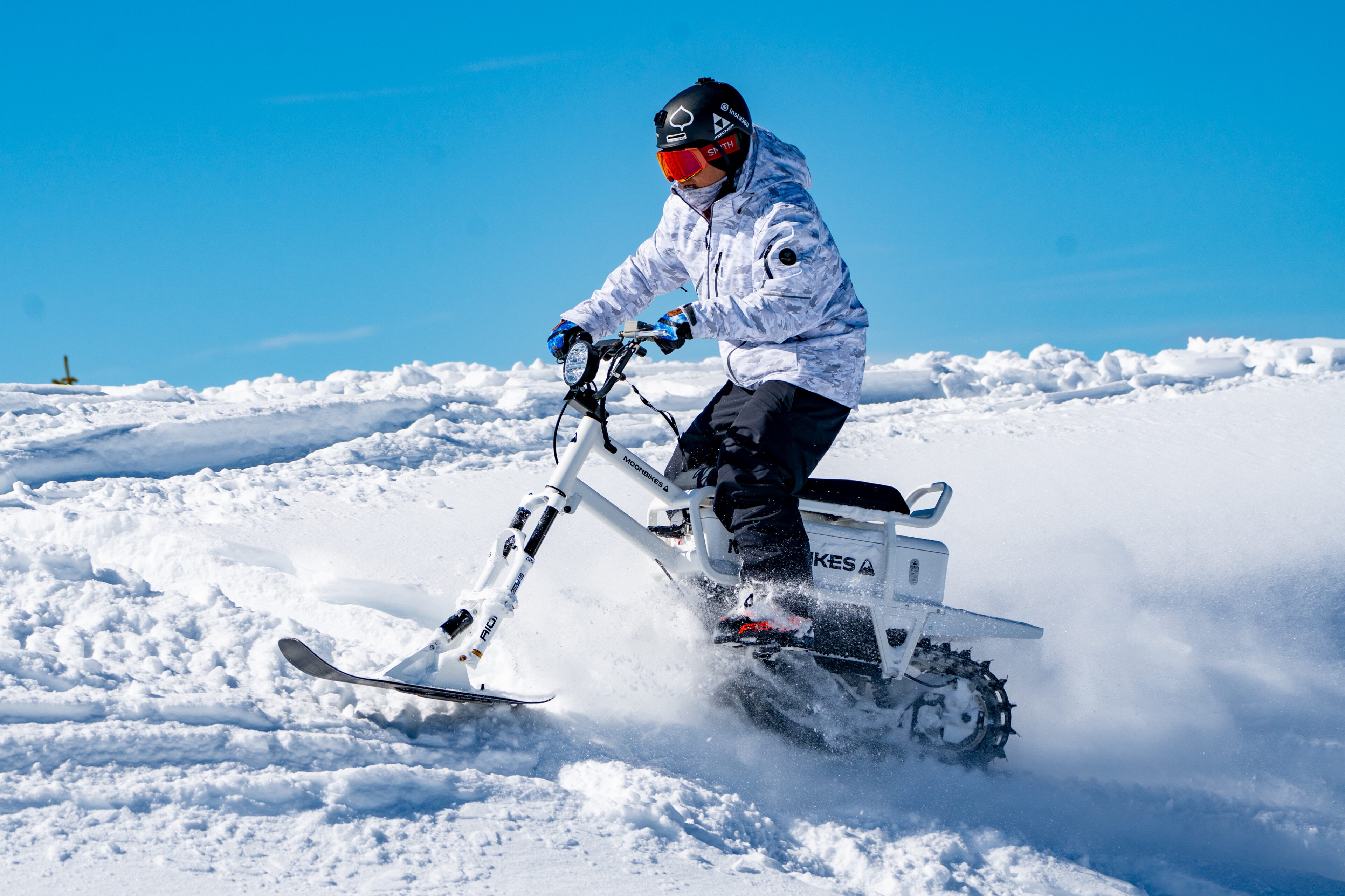 A person rides a snow bike over a fresh blanket of snow under a clear blue sky, wearing winter gear and a red helmet.