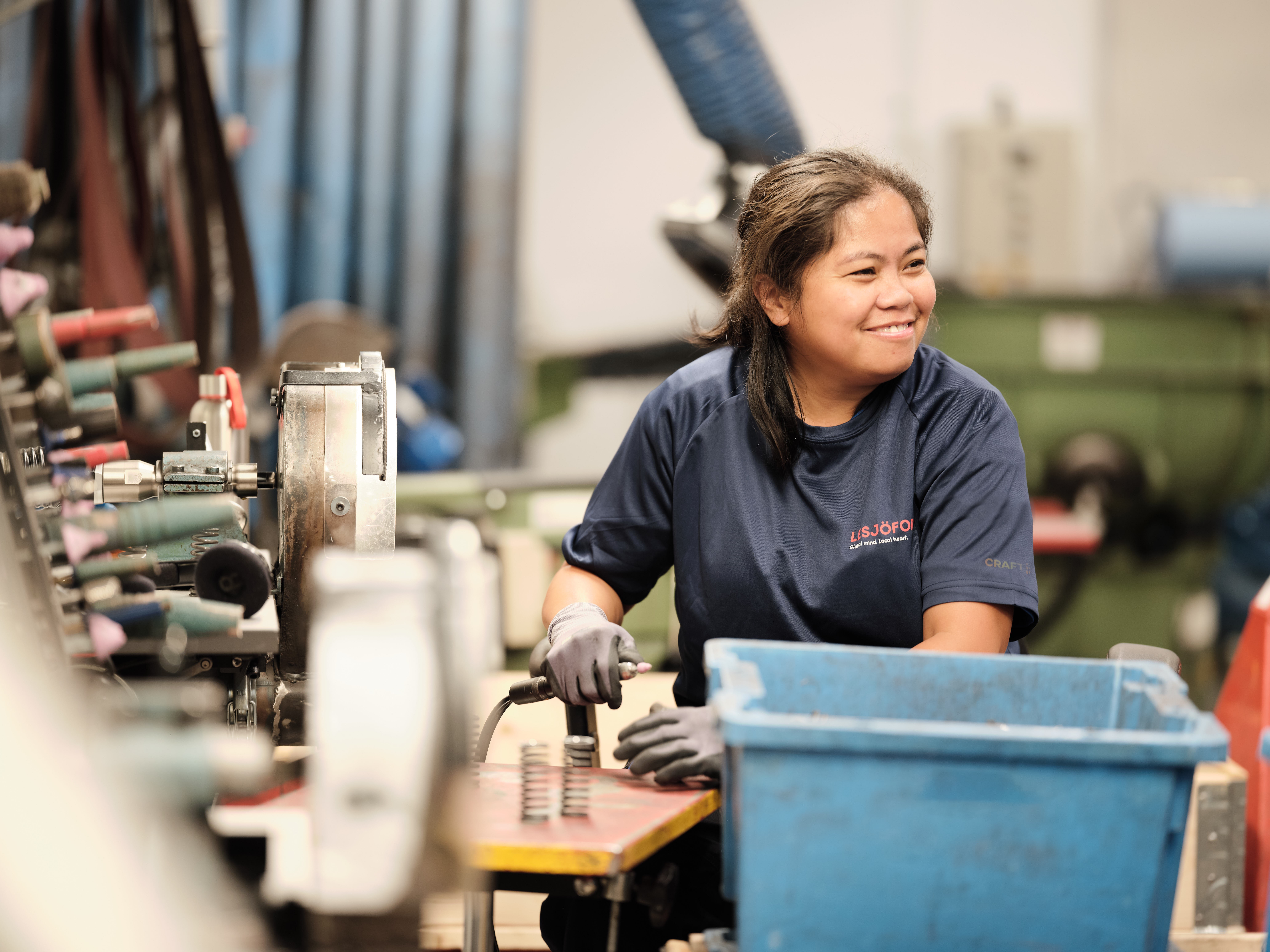 A woman in a navy shirt works happily at a cluttered workbench, surrounded by tools and springs, her focus directed to the right, in a busy workshop.