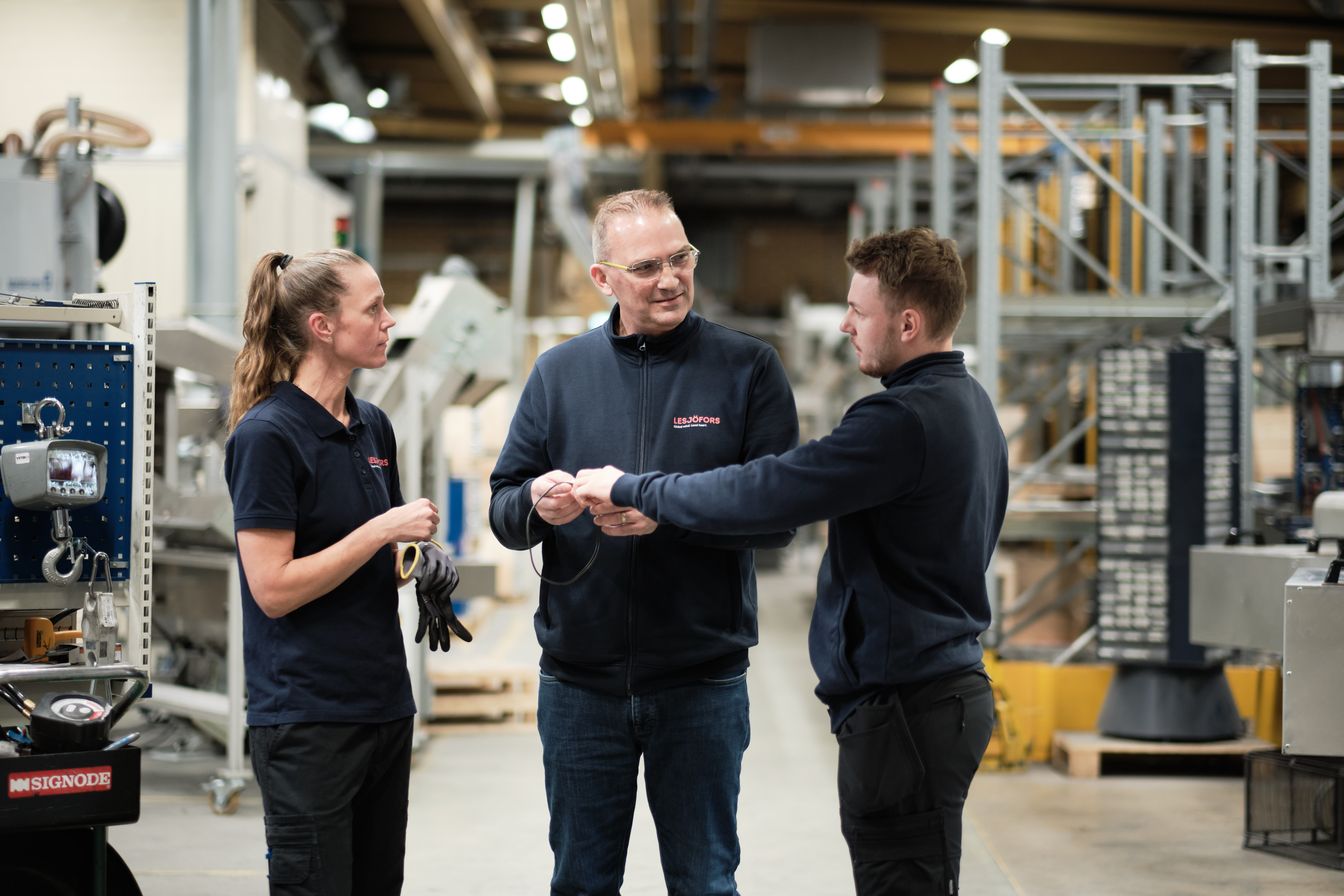 Three people in a factory setting, two young individuals listening attentively to an older person. Machinery and equipment are visible in the background.