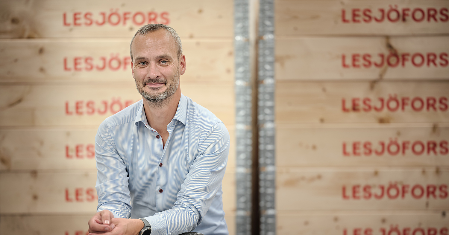 A man in a light blue shirt smiles while sitting in front of wooden crates marked "Lesjöfors."