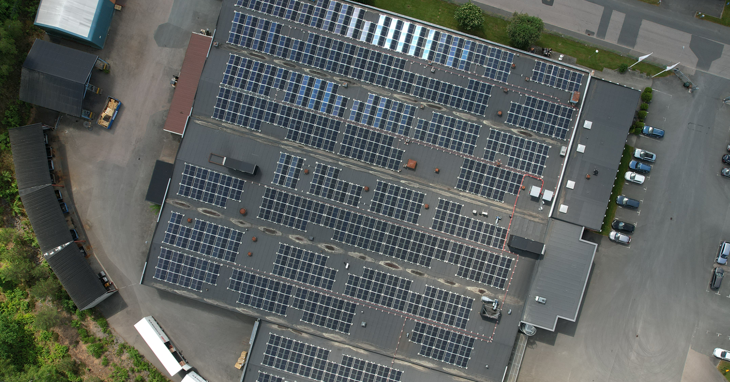 Aerial view of a large industrial rooftop covered with numerous solar panels, surrounded by greenery and parking areas.
