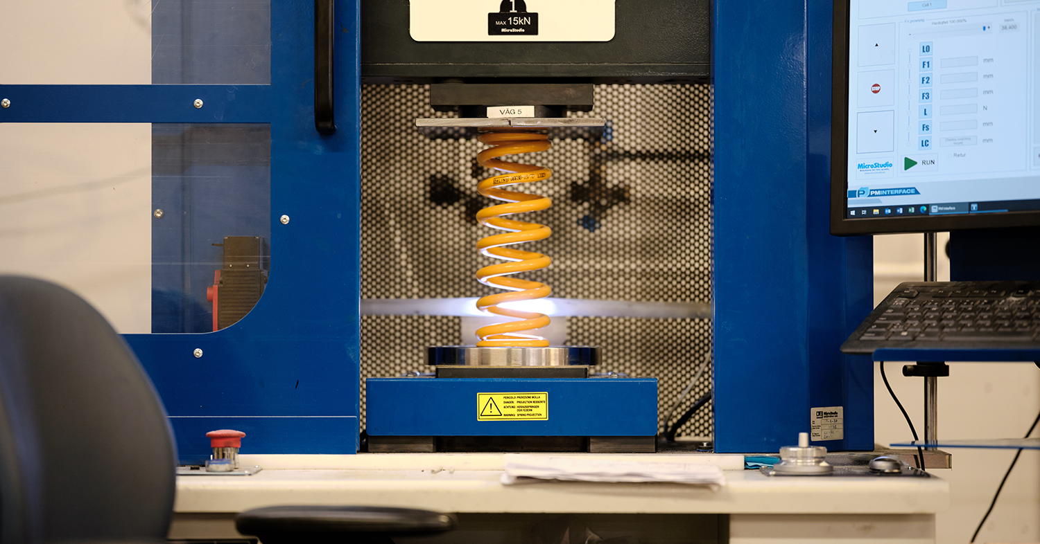 Laboratory setup showing a spring compression test. A gold-colored spring is being compressed by a machine, with a computer visible to the right.