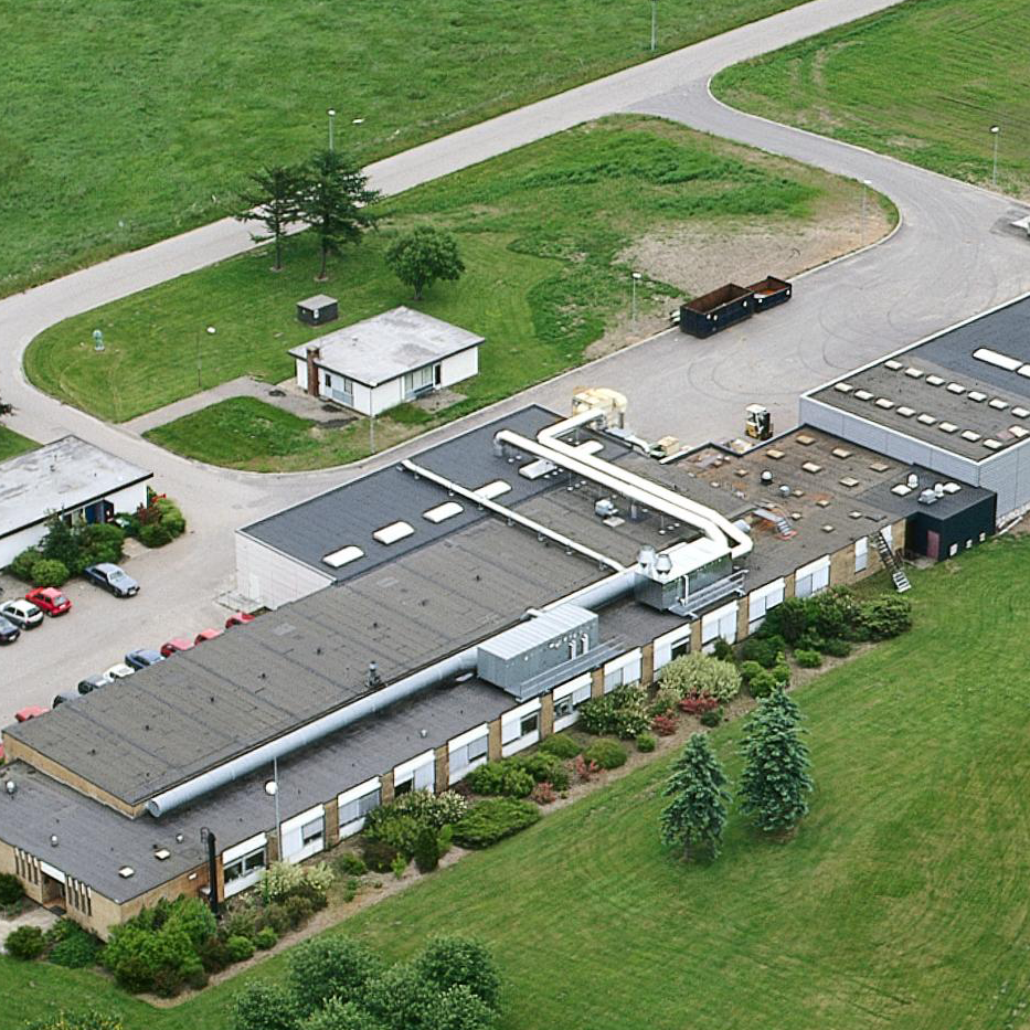 Aerial view of an industrial building with a flat roof, surrounded by green lawns and trees. A driveway curves around the building.
