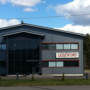A modern industrial building with "Lesjöfors" sign, large windows, and a sleek design. It's set against a backdrop of trees under a partly cloudy sky.